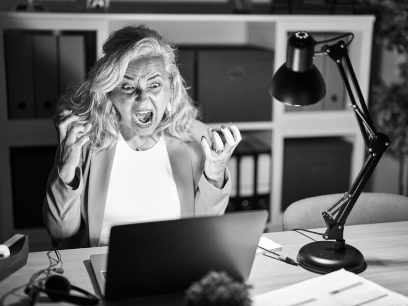 Middle age woman with grey hair working using computer laptop late at night crazy and mad shouting and yelling with aggressive expression and arms raised. frustration concept.
