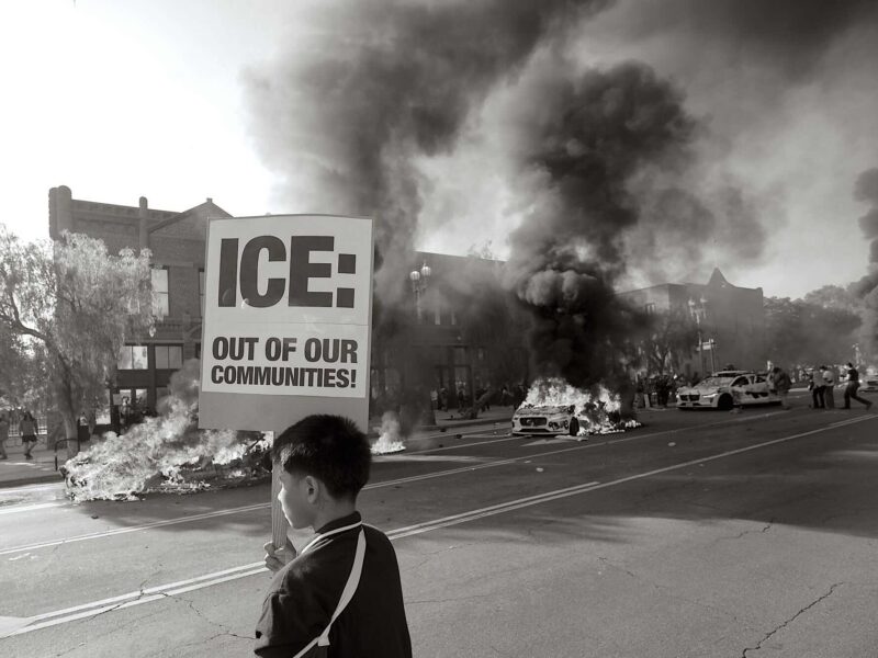Child holding anti ICE sign at protest in front of burning electronic vehicles